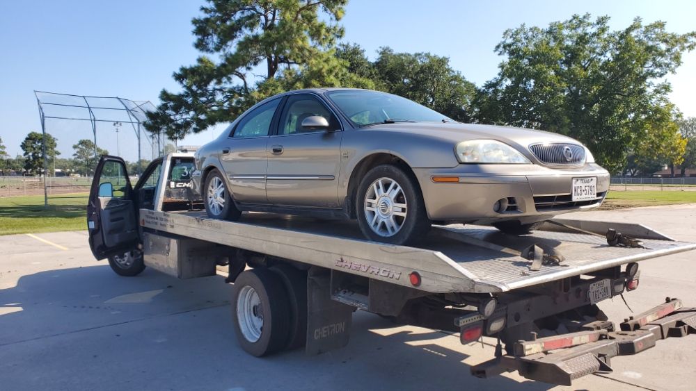 A silver sedan is loaded onto a tow truck in a parking lot near a baseball field, showcasing the vehicle's transport for repairs or towing.